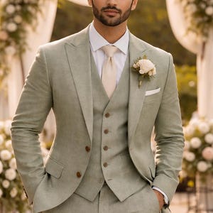 May include: A light green three-piece suit with a white shirt and cream tie. The suit features a two-button jacket, waistcoat, and trousers. A flower boutonniere is pinned to the lapel. The man is standing outside.