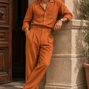 May include: A burnt orange long-sleeved button-down shirt with a collar and a chest pocket, paired with matching high-waisted pleated trousers. The outfit is worn with dark brown leather loafers and a wristwatch with a brown leather strap.