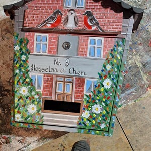 Può includere: Cassetta delle lettere dipinta a mano con un design a facciata in mattoni, con tre uccelli appollaiati sul tetto. La cassetta delle lettere ha la forma di una casa, con il testo "Nr. 9 Hesselaa & Chen" sul davanti ed è decorata con elementi floreali.