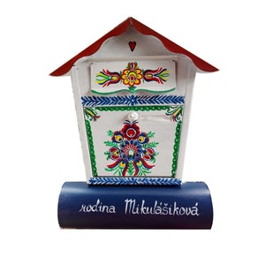 May include: White wooden mailbox with a red roof and a blue base. The mailbox is decorated with colorful folk art flowers and the text "rodina Mikulášiková".