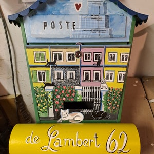 Può includere: Cassetta delle lettere dipinta a mano con tetto blu e bordi verdi. La parte anteriore presenta edifici colorati con gatti e la parola "POSTE". Un cartello giallo sottostante recita "de Lambert 62".