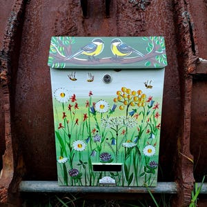 May include: A hand-painted mailbox with a nature theme. The top features two birds on a branch, while the front depicts wildflowers and insects. The color palette includes green, white, yellow, and red.