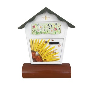 Könnte beinhalten: Weißer Holzbriefkasten mit grünem Dach und Sonnenblumenmotiv. Der Briefkasten hat eine kleine Klappe für die Post und einen braunen Holzsockel.