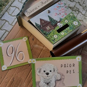 May include: A set of three hand-painted wooden signs. The first sign is a house number plaque with the number 96 in white on a beige background. The second sign is a warning sign with the text "POZOR PES" in brown on a beige background. The third sign is a mailbox with a painted scene of a house with flowers and a bird. The mailbox is painted in shades of brown, green, and white.