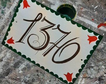 Può includere: Un cartello bianco con un bordo verde a festoni e tulipani rossi dipinti su ogni angolo. Il numero 1370 è scritto in corsivo nero sul cartello.