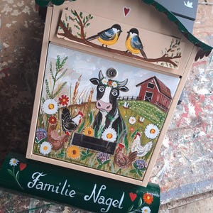 Op de afbeelding: Een beige metalen brievenbus met een handgeschilderd boerderijtafereel met een koe, kippen, bloemen en een rode schuur. De bovenste klep toont twee vogels op een tak. Een donkergroene cilindrische krantenhouder onderaan toont de tekst Familie Nagel.