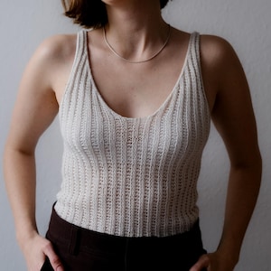 Könnte beinhalten: Ein hellbeige gestricktes Tanktop mit V-Ausschnitt. Das Oberteil hat eine gerippte Textur und wird mit einer dunkelbraunen Hose kombiniert. Eine silberne Halskette wird um den Hals getragen.