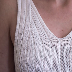 Könnte beinhalten: Nahaufnahme eines cremefarbenen, gerippten Strick-Tanktops mit V-Ausschnitt. Der Stoff hat ein strukturiertes Aussehen mit vertikalen Rippen. Das Tanktop ist ein einfaches, ärmelloses Kleidungsstück.