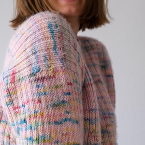 Könnte beinhalten: Ein hellrosa Strickpullover mit Rundhalsausschnitt und langen Ärmeln. Der Pullover hat ein gesprenkeltes Muster mit blauen, gelben und roten Akzenten. Das Strickwarenmaterial wirkt weich und bequem.