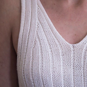 May include: Cream-colored knitted tank top with a V-neck. The ribbed texture creates vertical lines, adding visual interest to the garment. The straps are wide, and the overall design is simple and elegant.
