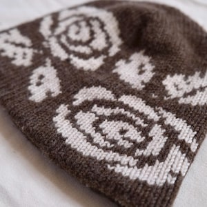 Könnte beinhalten: Eine dunkelbraune Strickmütze mit einem kontrastierenden cremefarbenen Blumenmuster. Die Mütze ist mit einem gerippten Rand und einem dichten, strukturierten Strickstich gefertigt und zeigt zwei markante rosenartige Motive auf der Vorderseite.
