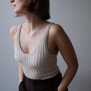 Puede incluir: Una camiseta sin mangas de punto acanalado color crema con cuello en V y tirantes finos. La parte superior se combina con pantalones marrones oscuros. Se ve un delicado collar plateado. La persona est&aacute; de pie frente a una pared gris claro.