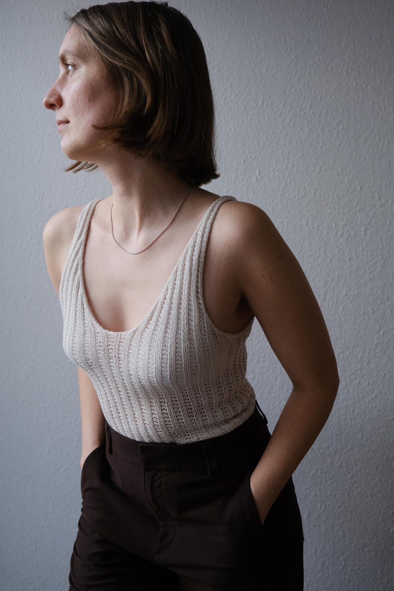 Puede incluir: Una camiseta sin mangas de punto acanalado color crema con cuello en V y tirantes finos. La parte superior se combina con pantalones marrones oscuros. Se ve un delicado collar plateado. La persona est&aacute; de pie frente a una pared gris claro.