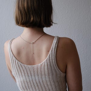 Puede incluir: Una camiseta sin mangas de punto acanalado color crema con cuello redondo y un delicado collar de plata con un peque&ntilde;o colgante. La modelo est&aacute; de espaldas a la c&aacute;mara, con el pelo corto casta&ntilde;o.