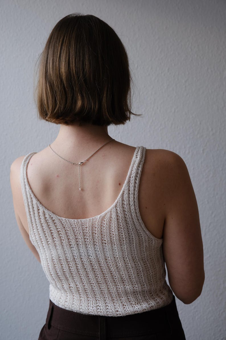 Puede incluir: Una camiseta sin mangas de punto acanalado color crema con cuello redondo y un delicado collar de plata con un peque&ntilde;o colgante. La modelo est&aacute; de espaldas a la c&aacute;mara, con el pelo corto casta&ntilde;o.