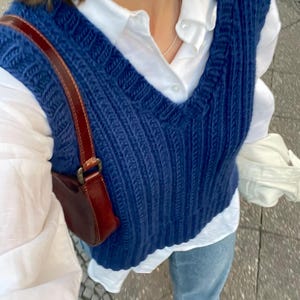 Könnte beinhalten: Eine marineblaue, gerippte Strickweste mit V-Ausschnitt, die über einem weißen Hemd getragen wird. Eine kleine, braune Lederhandtasche mit Schulterriemen ist sichtbar. Das Outfit wird durch hellblaue Jeans vervollständigt.