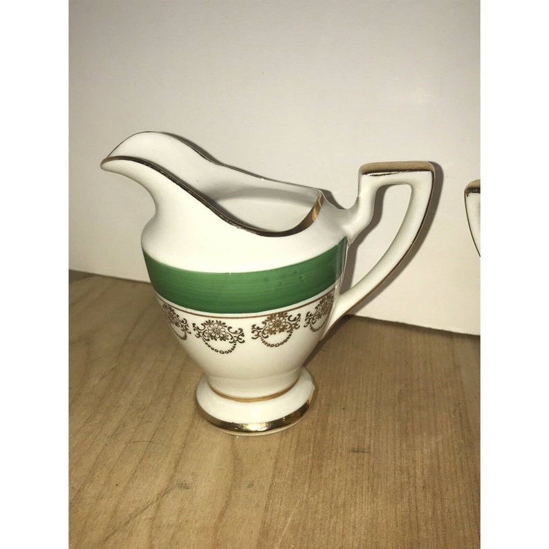 Puede incluir: Una jarra de crema blanca con una banda verde y ribetes dorados. La jarra tiene un delicado dise&ntilde;o floral y un borde dorado.