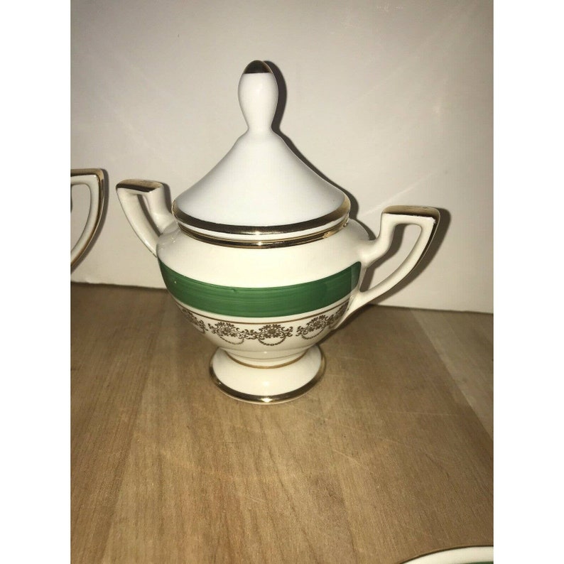 Puede incluir: Azucarero de porcelana blanca con una banda verde y ribete dorado. La tapa tiene un pomo en la parte superior.