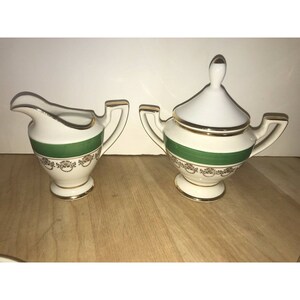 Puede incluir: Un juego de crema y az&uacute;car blanco con ribetes dorados y una banda verde. El juego est&aacute; sobre una superficie de madera.