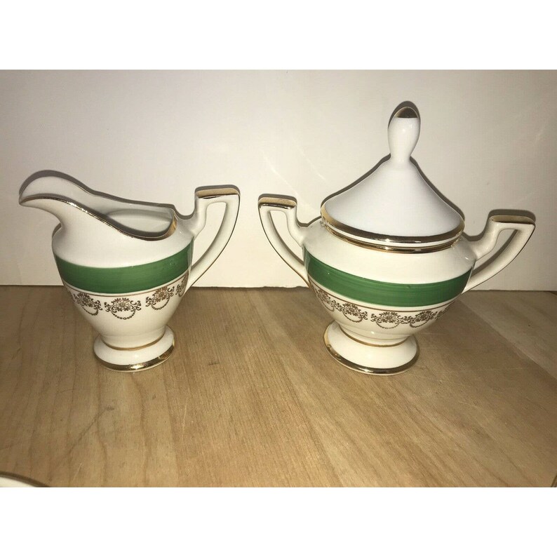 Puede incluir: Un juego de crema y az&uacute;car blanco con ribetes dorados y una banda verde. El juego est&aacute; sobre una superficie de madera.