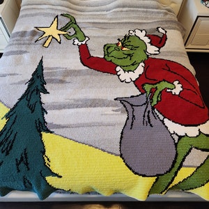 May include: A crocheted blanket featuring the Grinch from the popular Christmas movie "How the Grinch Stole Christmas". The Grinch is depicted in a red and white suit, holding a bag and reaching for a star. The blanket is a light gray with a yellow and green background.