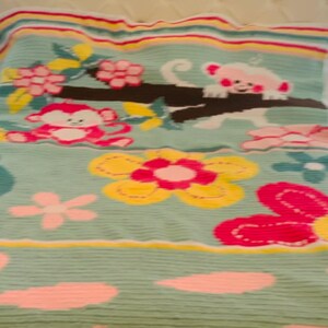 Könnte beinhalten: Eine hellblaue Bettdecke mit einem sich wiederholenden Muster aus rosa und weißen Affen, Blumen und Blättern. Die Bettdecke hat einen gestreiften Rand in Gelb-, Rosa- und Grüntönen. Die Bettdecke liegt auf einem Bett mit cremefarbenem Kopfteil.