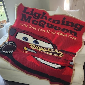 Könnte beinhalten: Eine handgefertigte rote Häkeldecke mit Lightning McQueen aus dem Film Cars. Die Decke trägt den Text "Lightning McQueen" und "2006 One Of A-Kind Race Car". Die Decke hat einen beigen Rand.