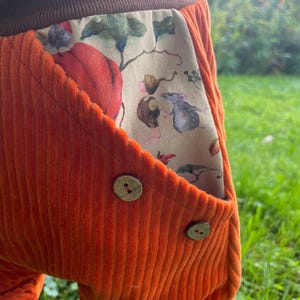 Könnte beinhalten: Orangefarbene Cordhose mit braunem Bund und einer Tasche mit Blumendruck. Die Tasche hat ein kleines Maus- und Kürbisdesign. Die Hose hat zwei Holzknöpfe.