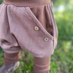 Könnte beinhalten: Braune Babyhose mit Fischgrätenmuster auf den Taschen und einem einfarbigen braunen Bund und Bündchen. Die Hose hat zwei Holzknöpfe an der Tasche und ist aus weichem Stoff gefertigt.