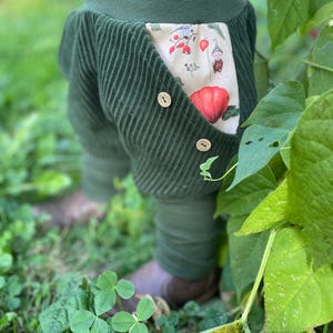 Könnte beinhalten: Grüne Cordhose mit einer aufgesetzten Tasche mit einem Blumendruck mit roten Beeren und einem kleinen Kobold. Die Hose hat zwei braune Knöpfe.