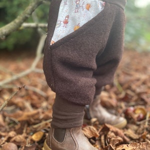 Könnte beinhalten: Braune Fleecehose mit einer großen Tasche an der Seite. Die Tasche ist mit einem weißen Stoff gefüttert, der ein Muster aus kleinen Figuren und Blättern zeigt. Die Hose wird von einem Kind getragen, das auf einem Bett aus braunen Blättern steht.