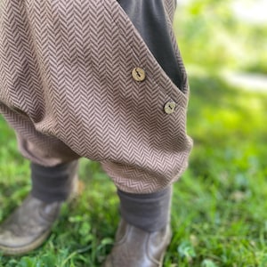 Könnte beinhalten: Braune und graue Hose mit Fischgrätenmuster und einem dunkelgrauen Bund und Bündchen. Die Hose hat zwei Taschen mit Holzknöpfen. Die Person trägt braune Lederstiefel und steht auf grünem Gras.