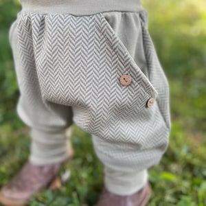 Könnte beinhalten: Salbeigrüne Babyhose mit Fischgrätenmuster auf den Taschen, befestigt mit Holzknöpfen. Die Hose hat einen bequemen, elastischen Bund und Bündchen. Das Kind trägt braune Lederschuhe.