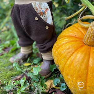Könnte beinhalten: Ein Kind trägt eine braune Fleecehose mit einer gemusterten Tasche und einem beigefarbenen Bund und steht neben einem großen orangefarbenen Kürbis. Die Hose hat zwei braune Knöpfe vorne. Der Kürbis liegt auf einem Bett aus grünem Moos und braunen Blättern.
