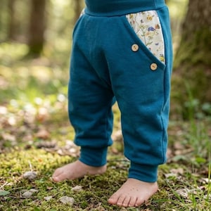 Könnte beinhalten: Tealfarbene Hose mit breitem Bund und Taschen mit einem cremefarbenen Blumen- und Schmetterlingsmuster. Die Taschenöffnungen sind auf jeder Seite mit zwei kleinen, runden, hellen Holzknöpfen verziert. Die Hose hat eng anliegende Bündchen an den Knöcheln.