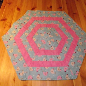 Könnte beinhalten: Ein sechseckiger Tischläufer mit einem blauen und weißen Muster aus Vögeln und einem rosa Rand. Der Läufer hat drei konzentrische rosa Bänder.