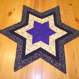 Könnte beinhalten: Ein gesteppter sternförmiger Tischläufer mit einem mehrschichtigen Design. Die Mitte ist ein tiefvioletter Stern, umgeben von einem cremefarbenen Stern mit goldenem Blumenmuster. Die äußere Schicht ist marineblau mit goldenen Sternen und Monden.