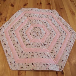 Könnte beinhalten: Ein handgefertigter, sechseckiger, gesteppter Tischset. Das Tischset hat ein Blumenmuster in Rosa-, Weiß- und Brauntönen. Breite rosa Bänder bilden ein geometrisches Design. Das Tischset liegt auf einem Holztisch.
