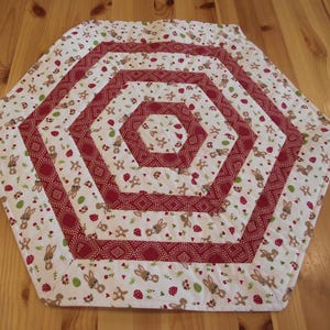 Könnte beinhalten: Ein sechseckiger Tischläufer mit Steppdesign. Das Muster zeigt Hasen, Blumen und Pilze auf weißem Grund. Die Ränder sind rot. Das Design ist symmetrisch. Die Maße sind in Zentimetern.