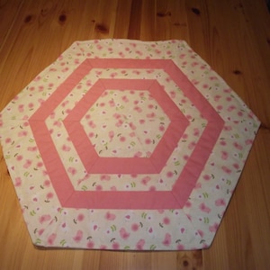 Könnte beinhalten: Ein sechseckiger Tischläufer mit weißem Hintergrund und rosafarbenem Blumenmuster. Der Läufer hat einen rosafarbenen Rand und drei konzentrische rosafarbene Ringe.