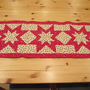 Könnte beinhalten: Ein roter und weißer Tischläufer mit einem Sternenmuster. Der Läufer ist aus Stoff mit einem Druck von Stechpalmenblättern.