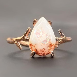 May include: A rose gold ring featuring a pear-shaped, white gemstone with pink and copper-colored inclusions. The ring's band is designed to resemble branches, adding a naturalistic touch to the jewelry.