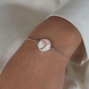 May include: A silver bracelet with a circular charm displaying a painted tree design. The charm has a white backdrop, a brown tree trunk, and pink blossoms. The bracelet is shown on a wrist.