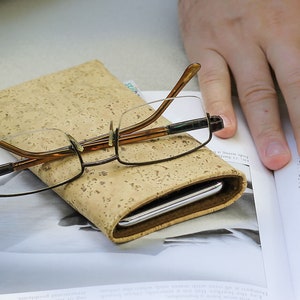 Puede incluir: Una funda cubierta de corcho con un marco marr&oacute;n y un tel&eacute;fono plateado en su interior. Un par de gafas con patillas marrones y lentes transparentes descansa sobre la funda.