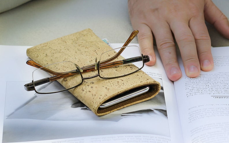 Puede incluir: Una funda cubierta de corcho con un marco marr&oacute;n y un tel&eacute;fono plateado en su interior. Un par de gafas con patillas marrones y lentes transparentes descansa sobre la funda.