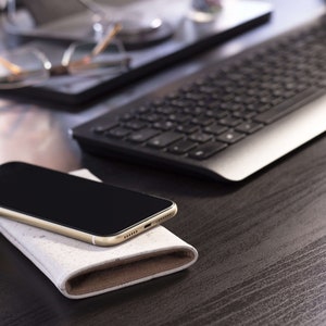 Puede incluir: Un tel&eacute;fono inteligente dorado con una pantalla negra descansa sobre una funda de cuero blanca y marr&oacute;n. El tel&eacute;fono est&aacute; sobre un escritorio de madera oscura con un teclado plateado en el fondo.