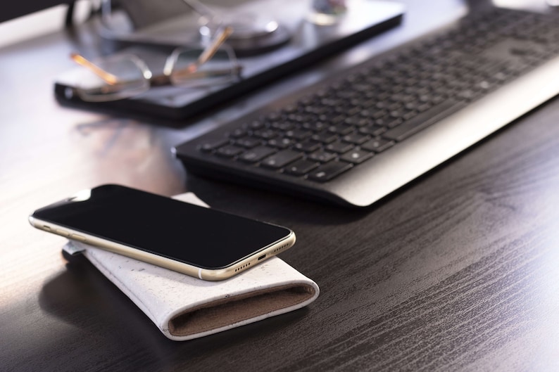 Puede incluir: Un tel&eacute;fono inteligente dorado con una pantalla negra descansa sobre una funda de cuero blanca y marr&oacute;n. El tel&eacute;fono est&aacute; sobre un escritorio de madera oscura con un teclado plateado en el fondo.