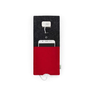 Op de afbeelding: Een grijze telefoonoplader van vilt met een rode viltzak voor een telefoon. Een witte telefoon zit in de zak en een witte oplaadkabel is aangesloten op de telefoon en de stekker.