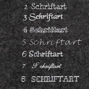 May include: A close-up of a black textured surface with nine examples of the word "Schriftart" written in different fonts and styles.