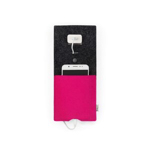 Puede incluir: Una estación de carga para teléfono de fieltro gris con un bolsillo de fieltro rosa para un teléfono y un cable de carga blanco. La estación de carga tiene un pequeño adaptador de corriente blanco y un botón negro.
